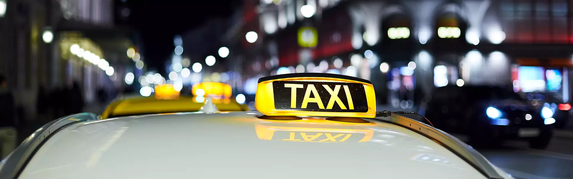 Ein Taxi mit einem beleuchteten „TAXI“-Schild auf dem Dach parkt nachts auf einer Stadtstraße in Erding, im Hintergrund sind verschwommene Lichter und Gebäude zu sehen.