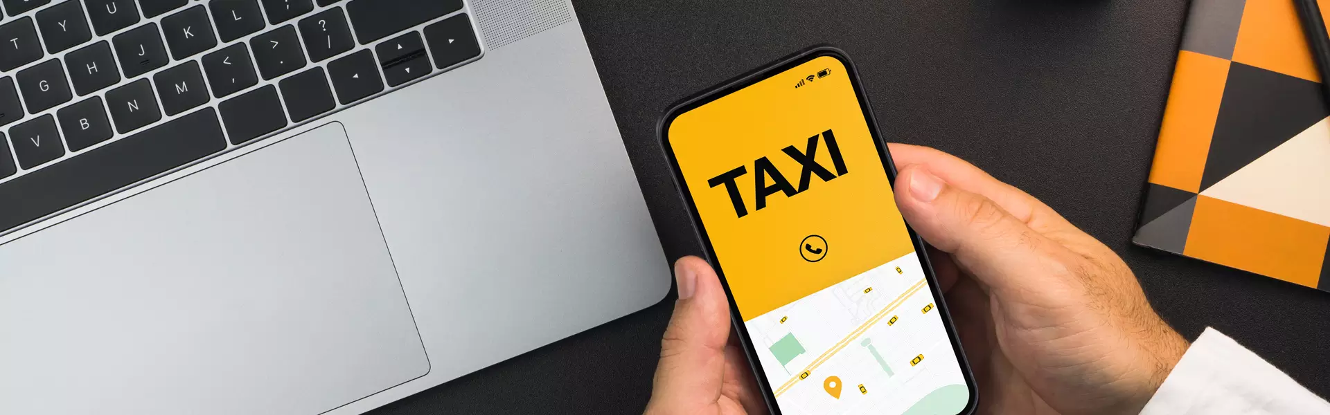 Eine Person hält ein Smartphone in der Hand und zeigt die Oberfläche einer Taxi-Buchungs-App an, auf der praktischerweise Taxidienste in Erding angezeigt werden. Neben dem Telefon liegen ein Laptop und ein buntes Notizbuch auf der dunklen Oberfläche.